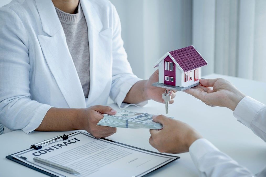 Hands exchanging a model house and keys over a stack of cash, with a contract on the table, illustrating a real estate transaction related to selling properties.